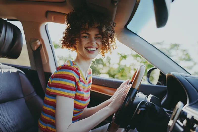 Young woman with curly hair sits in a car smiling as she drives, a candid lifestyle moment captured with retro film color and warm natural tones.