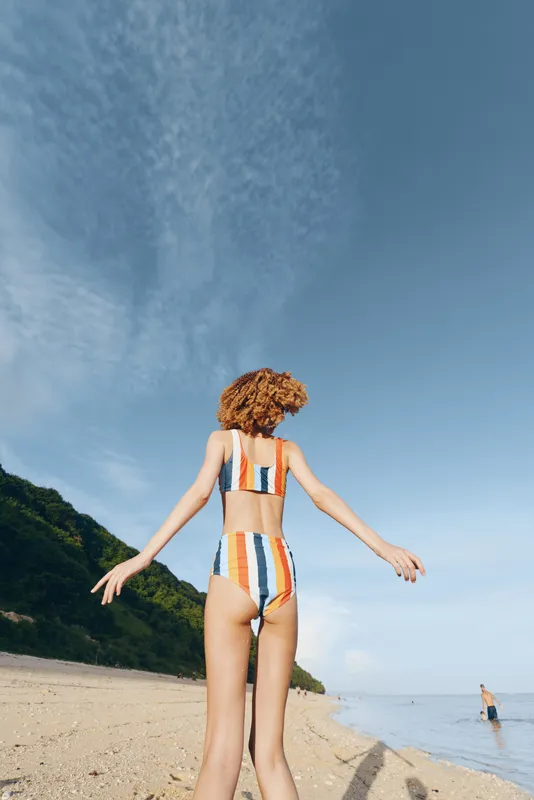 Woman in striped swimsuit walks on beach toward ocean waves, summer sunlight and blue sky, scenic coastline with hills, back view, carefree vacation mood, travel lifestyle