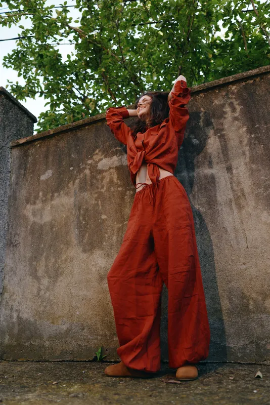 Woman in a bold orange jumpsuit stands against a weathered wall outdoors, showcasing confident street fashion and a playful pose. Bright colors and natural light create a dynamic fashion mood