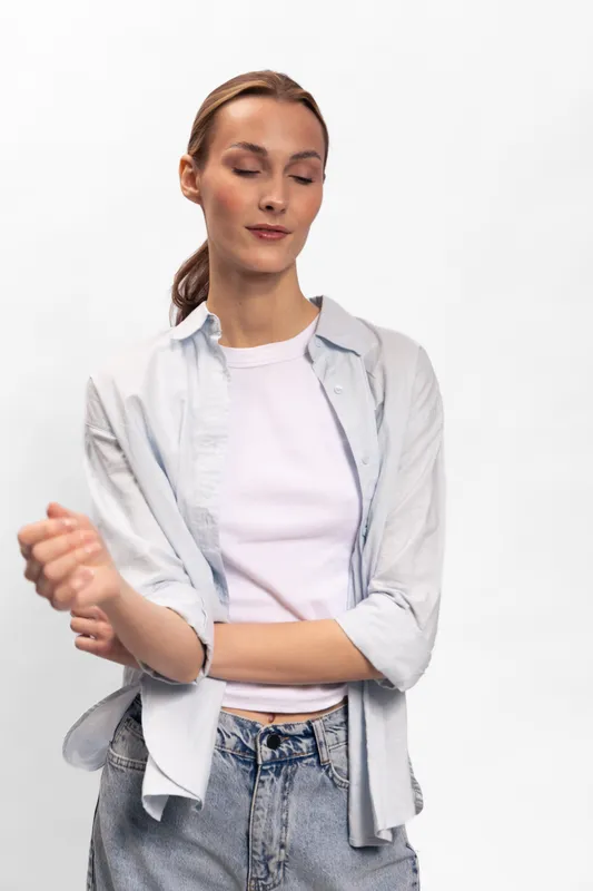 Young woman in casual clothing poses with eyes closed and relaxed expression, standing against a white background. Concept of calmness, peace, and casual fashion style.
