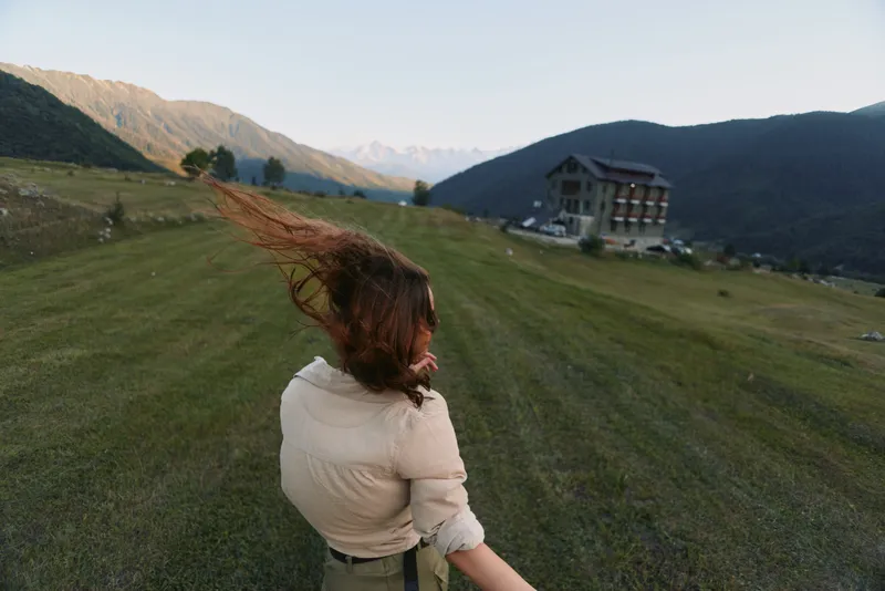 Woman outdoors, back view, wind in hair, sunlit hillside scene, casual lifestyle moment, expansive mountains, green field, peaceful vibe, natural colors, remote valley