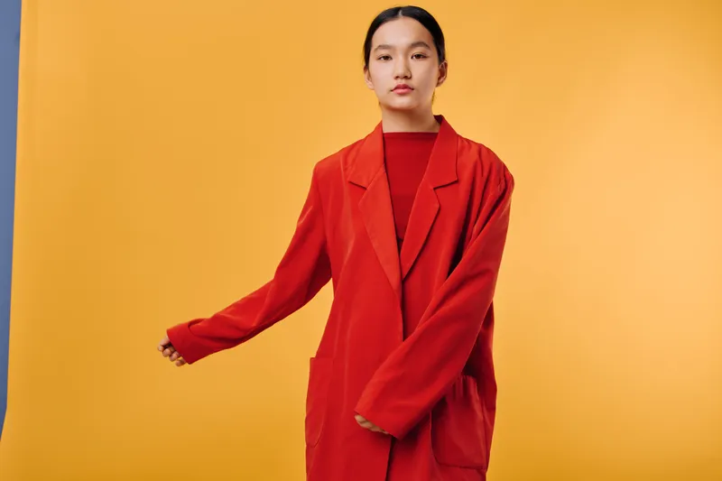 Portrait of young woman in red oversized blazer standing against yellow background with serious expression and natural makeup, fashion style with minimalistic studio setup.