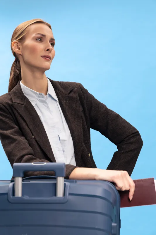 Confident woman in formal jacket holds blue suitcase and passport against clear blue background. Concept of business travel, professional woman, and modern lifestyle.