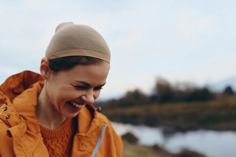 Woman outdoors, laughing in a knit hat and jacket by a lake, winter lifestyle, warm smile in sunlight, soft breeze, relaxed closeup, natural countryside background