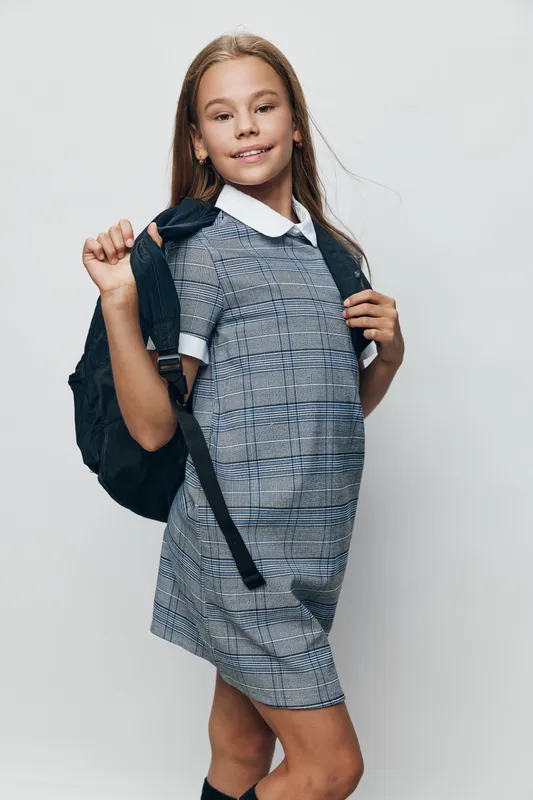 Girl child with long hair wearing school uniform dress and backpack standing with confident smile on white background in studio. Back to school readiness and education concept.