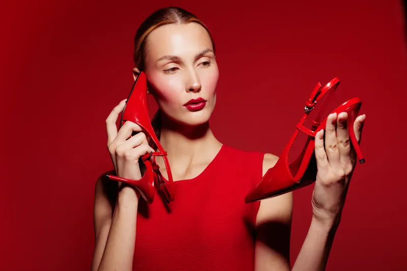 Woman in red dress holds red high heels near face on red background. Concept of fashion, style, and bold color contrast with elegant female model posing in studio lighting.
