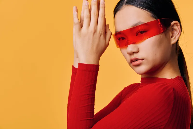 Young woman in red sweater and futuristic red glasses posing with palms pressed together on orange background. Closeup portrait with modern fashion and confident expression.