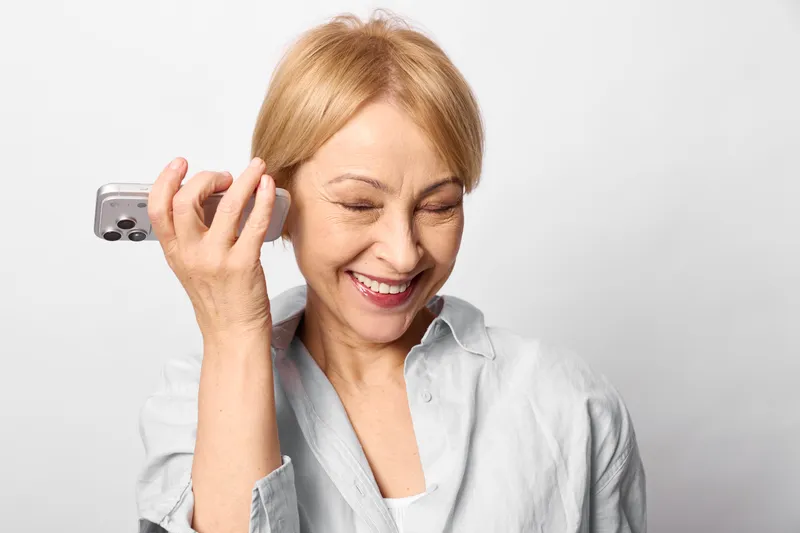 Elderly woman on a white background holds a smartphone phone to her ear, smiling while showing stress anxiety and headache, mental health emotional communication