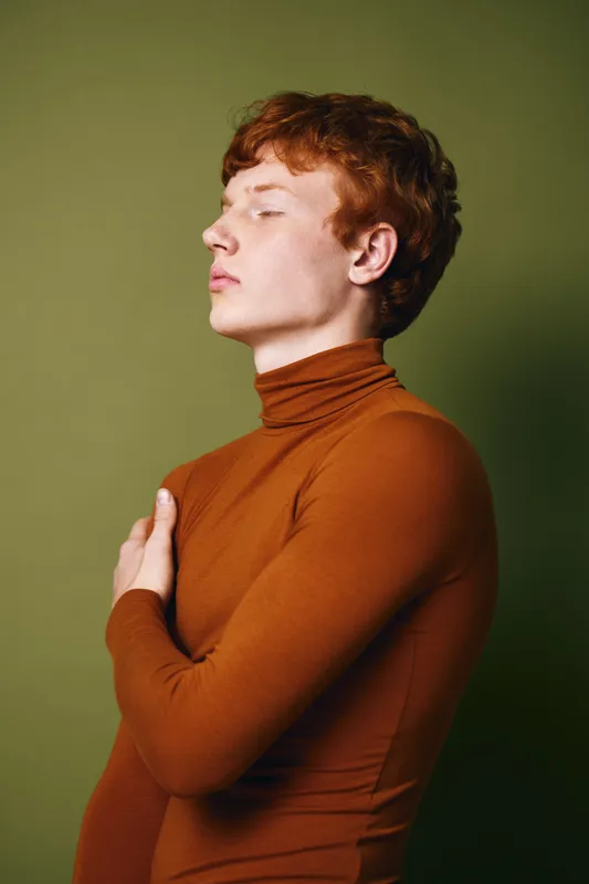 Portrait of young man with red hair wearing brown turtleneck looking sideways with closed eyes against green background. Fashion model posing with thoughtful expression in studio.