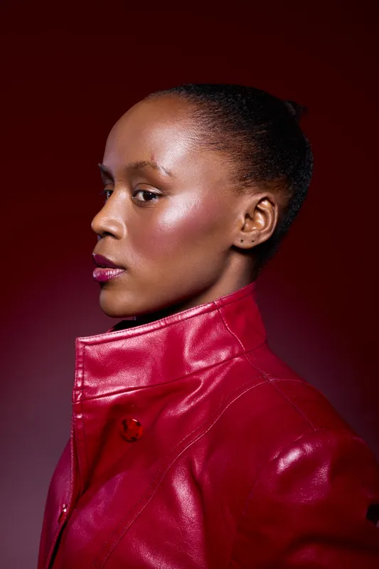 Profile portrait of an African woman with dark skin wearing a red leather jacket. The studio lighting highlights her smooth skin and confident expression on a dark red background.