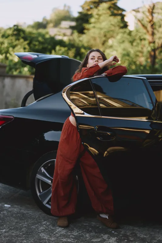 A stylish woman in a burnt orange jumpsuit leans against a sleek black car with its door open, capturing a candid lifestyle moment outdoors, suggesting independence and urban exploration