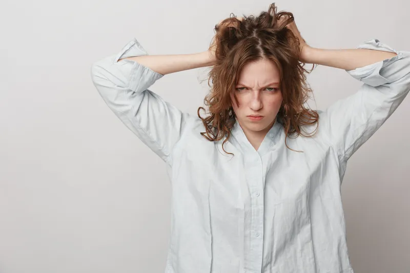 Frustrated young woman with curly hair wearing white shirt pulls her hair and frowns. Isolated on light background. Concept of stress, anger, frustration, and emotional tension.