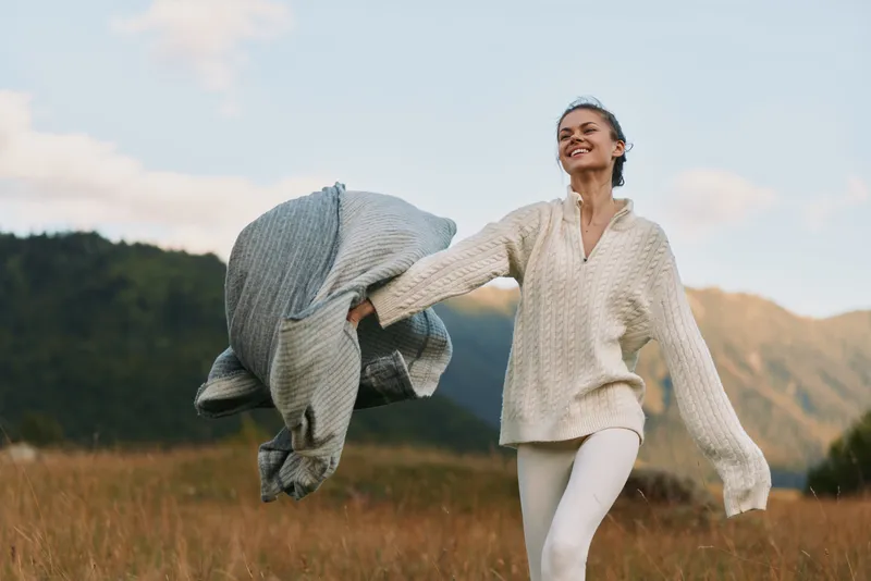 Young woman in cozy knitwear joyfully spins outdoors in a mountain meadow during autumn, holding a gray scarf with a carefree smile on her face.