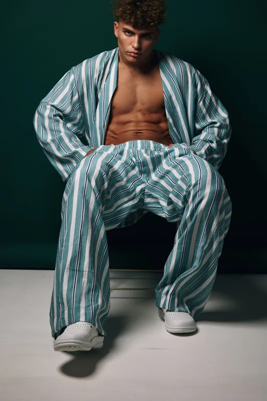 Young man sitting in striped pajama pants and opened matching shirt showing muscular torso and abs on dark background. Fashion model poses with confident expression seated in studio and casual