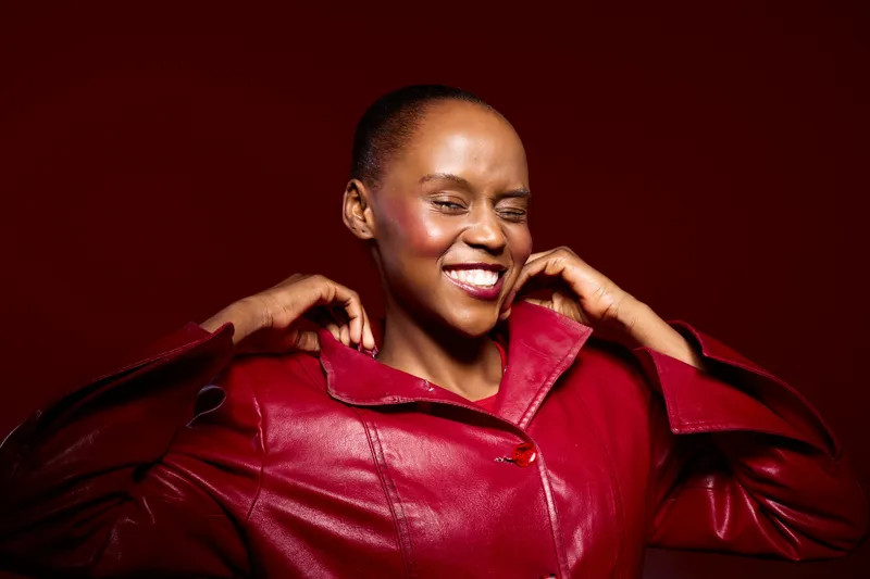 Confident black woman with dark skin smiles broadly while adjusting her vibrant red leather jacket against a deep red background. Concept of fashion, joy, and empowerment.