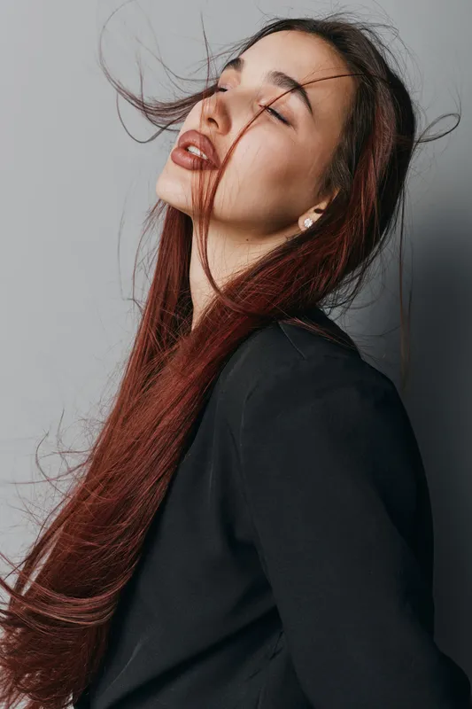 Woman with long red hair and closed eyes wearing black top posing with wind blowing hair in studio. Portrait of female model with natural makeup and serene expression on gray background.
