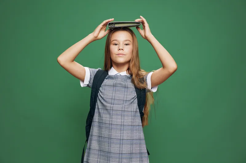 Girl child in dress holding book on head standing with backpack on green background for education, knowledge, learning and school concept of student growth and youth.