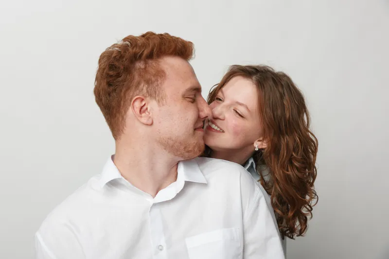 Happy young couple in white shirts embraces and smiles with closed eyes on light background. Concept of love, affection, relationship, and romantic moment between man and woman.
