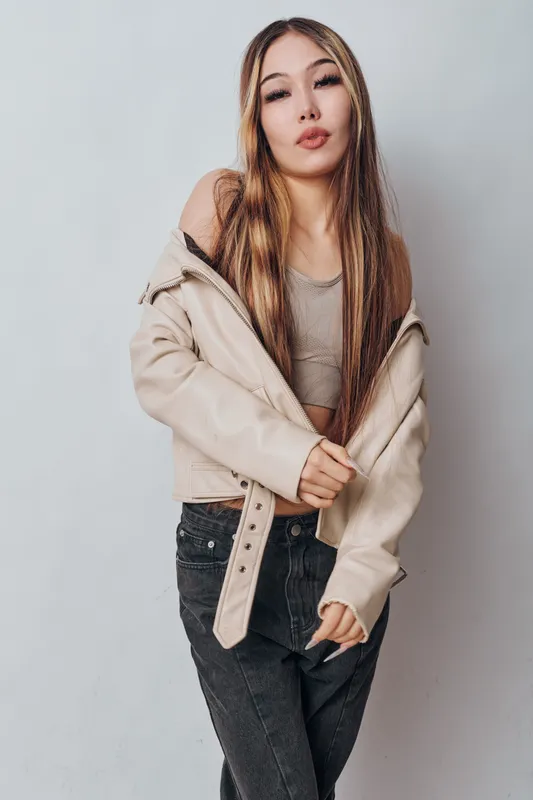 Young woman with long hair wearing beige jacket and grey top posing against neutral background. Fashion model standing with casual confident style and trendy outfit indoors.
