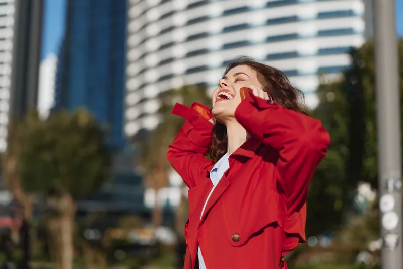 A joyful woman in a red coat smiles in bright outdoor light, embodying carefree confidence and urban street style against a modern city backdrop, sunny sky.