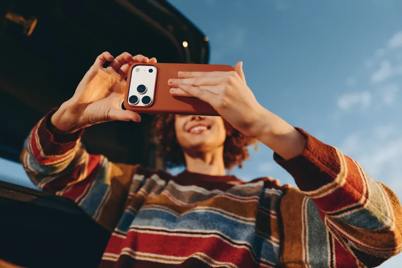 Woman holds a smartphone for a selfie, smiling outdoors with curly hair, striped sweater sleeves and hands framing the phone against a bright sky, lifestyle moment