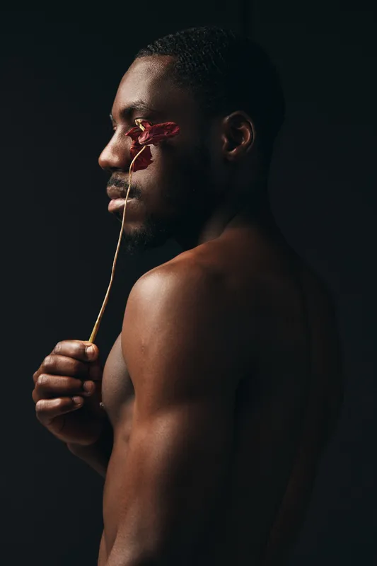 African man with dark skin holding dried flower near face in creative conceptual visual style. Artistic portrait with bare shoulders and thoughtful expression on black background.