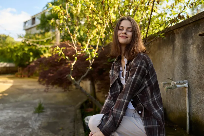 Smiling woman sitting outdoors in a plaid jacket, enjoying bright daylight. Casual lifestyle moment captured in a sunlit garden setting, emphasizing comfort, confidence, and simple happiness