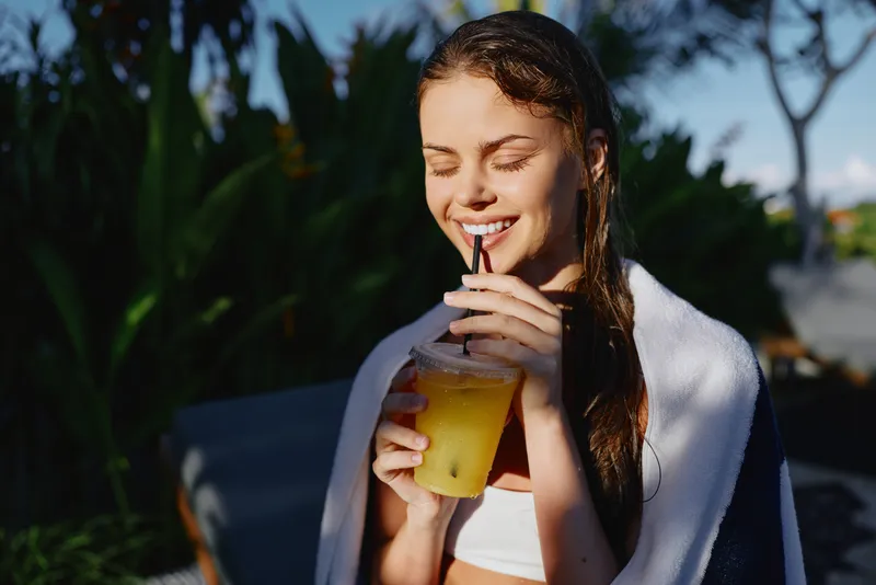 Bright outdoor lifestyle shot of a smiling young woman sipping lemonade, wrapped in a towel after sunbathing, relaxed casual vibe, warm daylight, natural setting with greenery and blue sky