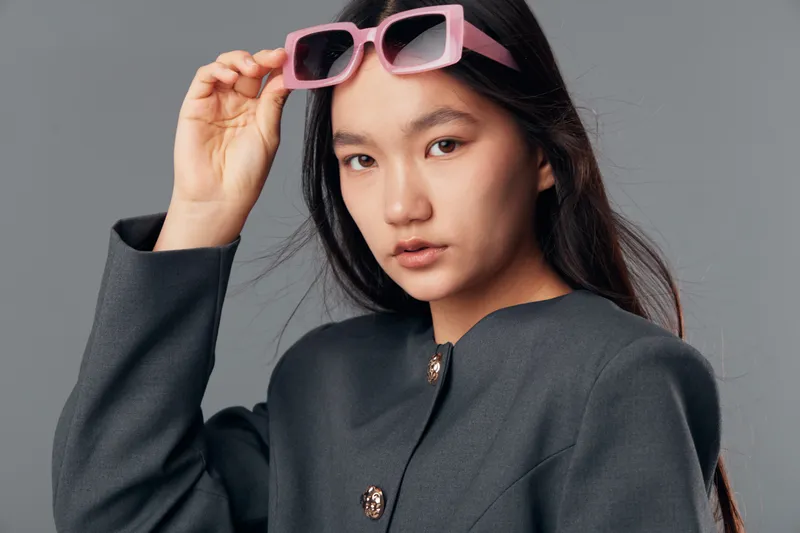 Young woman with long dark hair wearing pink sunglasses and a gray jacket looking at camera with serious expression on plain gray background fashion style portrait.