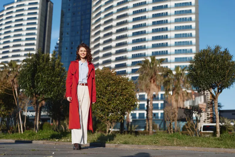 Cheerful woman outdoors wearing a bright red coat, standing on a sunny urban street with modern buildings, palm trees, and a relaxed, confident pose in soft daylight.