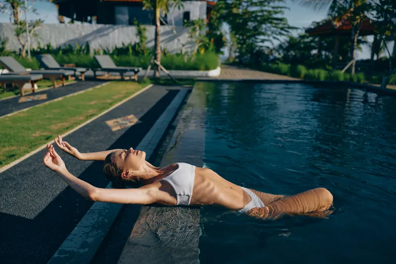 Young woman in white bikini relaxes floating on water at outdoor pool under sunlight with eyes closed enjoying summer lifestyle and peaceful moment near green garden.