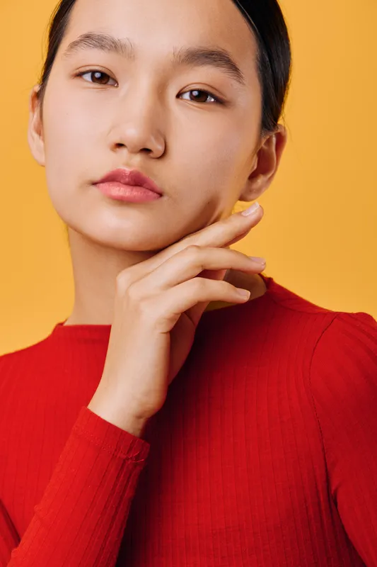 Woman portrait with hand near face in red sweater against yellow background. Closeup of young adult female with natural expression and smooth skin in studio environment.