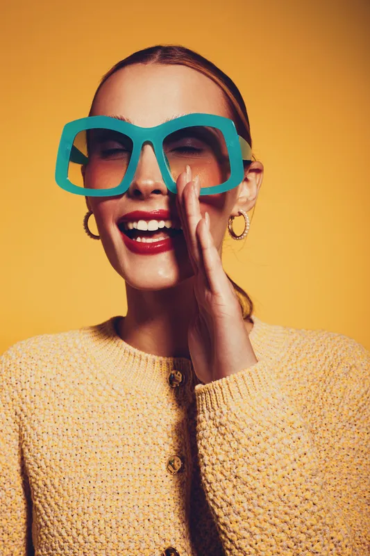 Smiling woman with large blue glasses and red lipstick laughs joyfully on yellow background. She wears a textured sweater and gold hoop earrings. Concept of happiness and fashion fun.