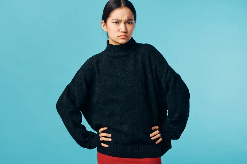 Young woman with dark hair and long sleeves wearing black sweater standing with hands on hips and serious expression on blue background in studio portrait.