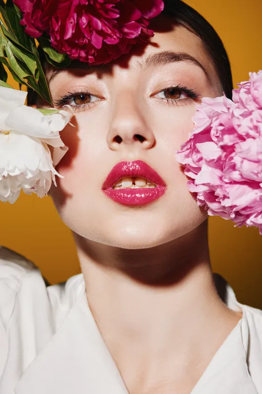 Close-up of young woman with glossy red lips poses between white and pink flowers on golden background. Creative beauty fashion concept with bright, smooth skin and delicate makeup.
