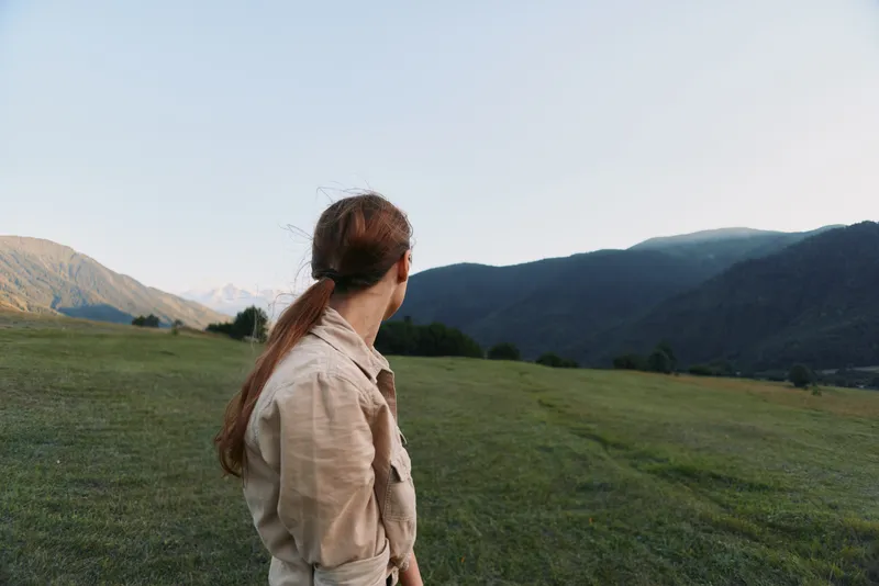 Woman seen from behind in a casual jacket stands in a lush field with distant mountains, creating a serene outdoors lifestyle scene focused on nature and exploration