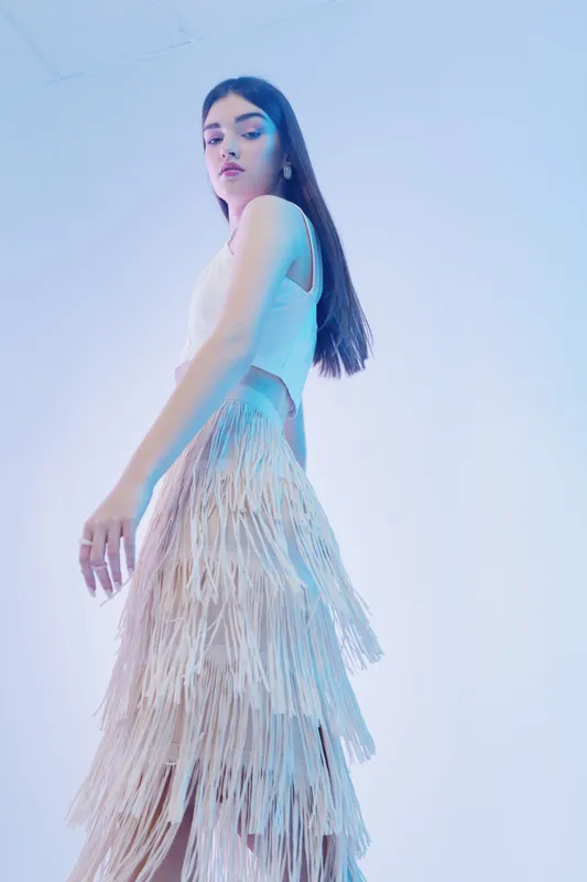 Young woman beauty fashion model poses in white fringed skirt and sleeveless top against light background. Concept of modern style, elegance, and confident expression.