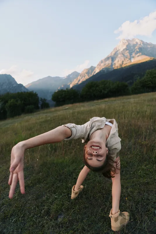 Happy woman outdoors doing backbend in grassy field with mountains in background, casual outfit, natural light, joyful moment, active lifestyle, summer day