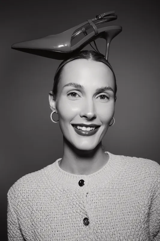 Smiling caucasian woman with light skin wearing hoop earrings and knitted sweater balances shiny high heel shoe on head in black and white portrait.