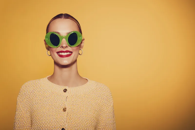Smiling woman with green round sunglasses and red lipstick wears beige knitted cardigan on yellow background. Concept of fashion, style, and cheerful mood in studio portrait.