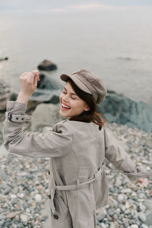 Happy young woman in beige trench coat and patterned cap smiling with closed eyes near rocky shore. Concept of freedom, joy, and casual autumn fashion by the sea on a cloudy day.