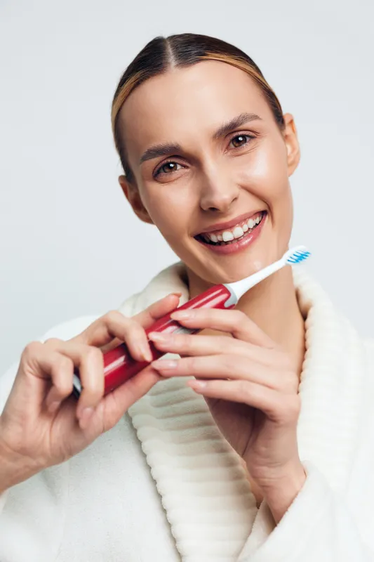 Smiling woman holding red electric toothbrush close to her face. White background with soft lighting. Concept of dental care, hygiene, oral health, and morning routine.