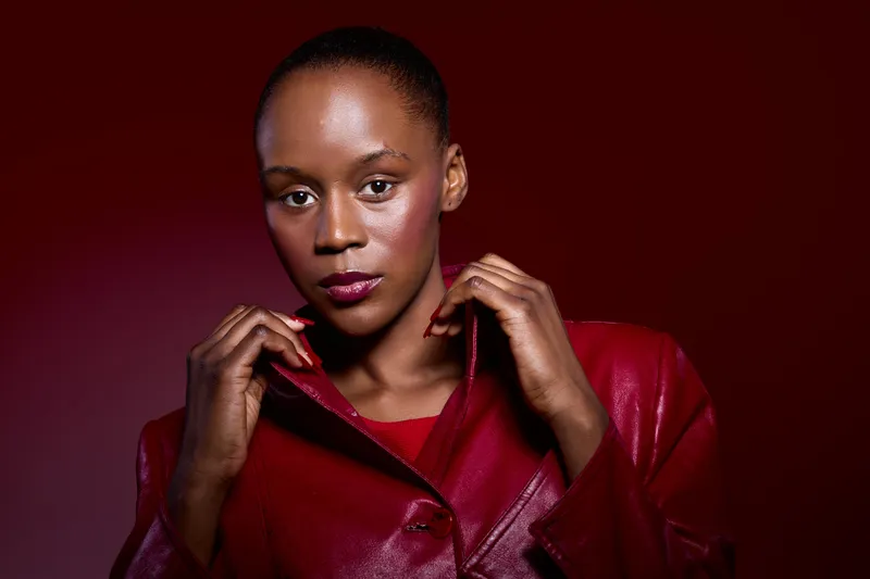 Confident black woman with dark skin wearing a red leather jacket poses against a red background. Studio portrait showcasing fashion, beauty, and elegance with strong lighting.