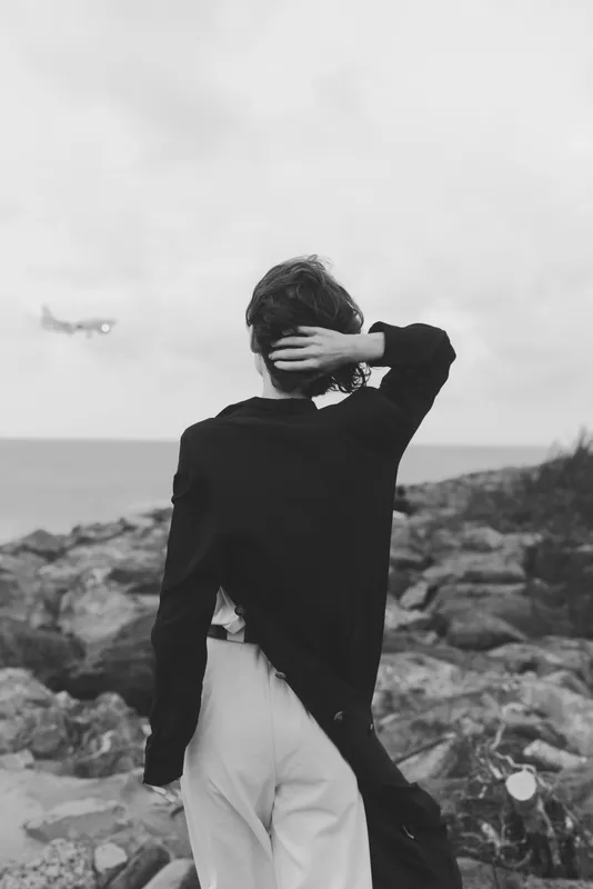 Silhouette, backview, fashion along a rocky coastline with ocean breeze, monochrome mood, coat draped, trousers, distant aircraft, reflective cinematic scene for travel or editorial use