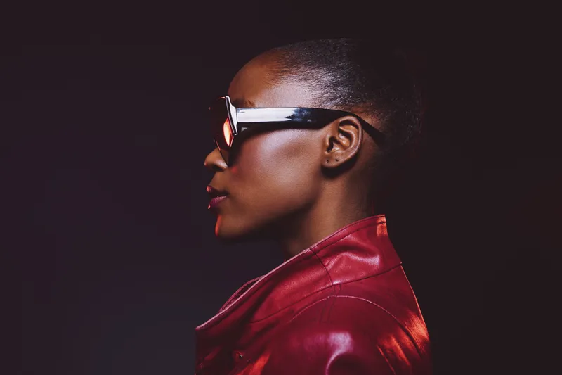 Profile portrait of a confident black woman with dark skin wearing stylish glasses and a red leather jacket against a dark background. Concept for fashion, empowerment, and modern style.