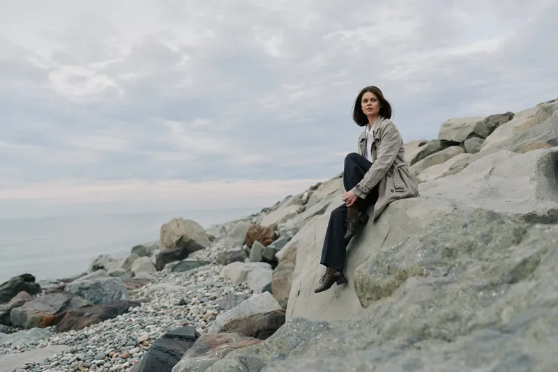A woman in a coat sits thoughtfully on large rocks by a pebble beach under a cloudy sky. The calm sea and natural landscape create a reflective, peaceful mood.
