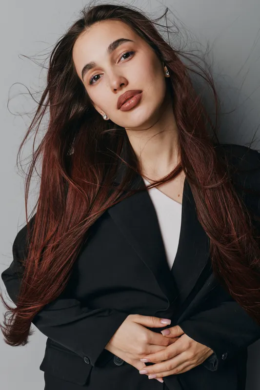 Young woman with long brown hair wearing black blazer posing against gray background. Portrait of confident female model with natural makeup and earrings in studio lighting environment.