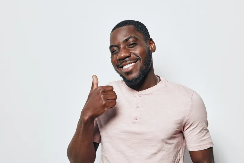 African American man with dark skin smiling and showing thumbs up gesture wearing casual light pink shirt on white background. Positive emotion and confident expression.