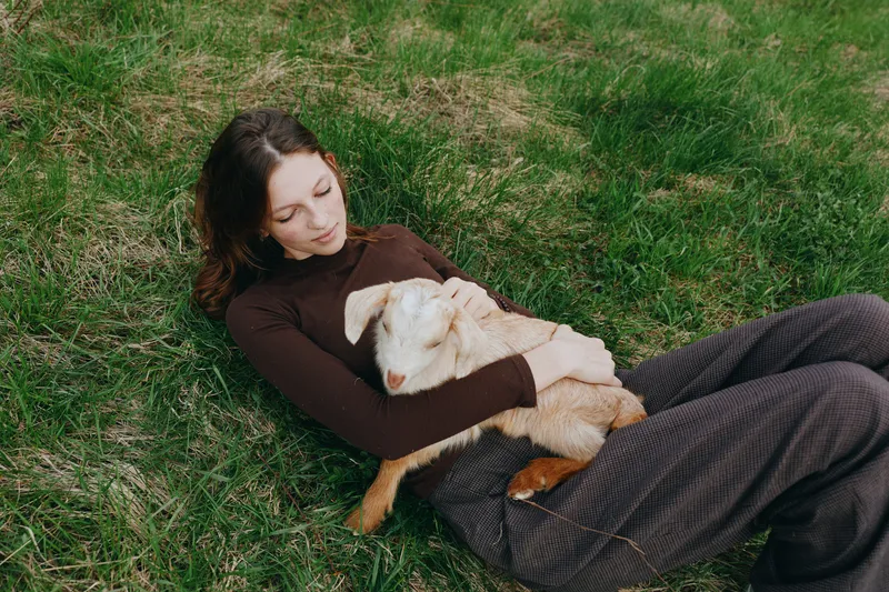 Outdoor scene of a woman lying on lush grass, gently cuddling a goat kid, sun warmed, cozy and relaxed, showcasing lifestyle, companionship, and a deep connection with nature