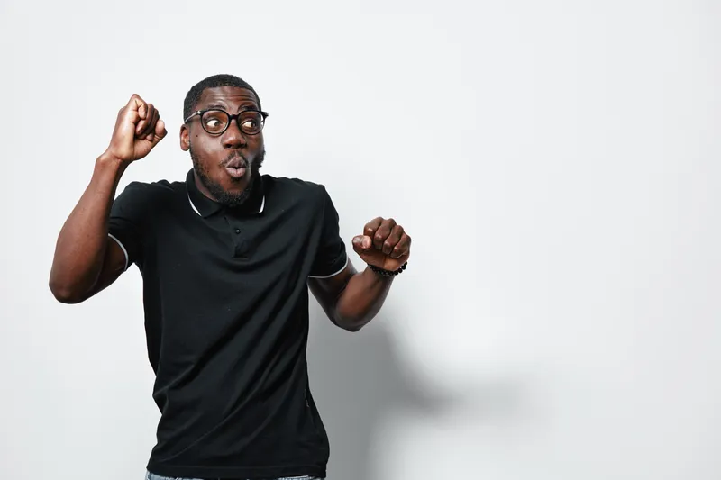 African American man with dark skin and glasses wearing black polo shirt showing surprised expression with raised fists on white background. Casual style and emotion concept.